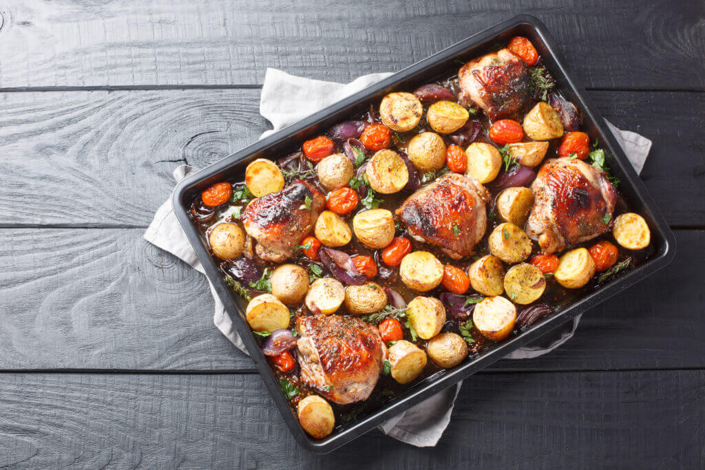 Chicken thighs baked with potatoes, garlic, onions and tomatoes close-up on a black baking sheet on a wooden table
